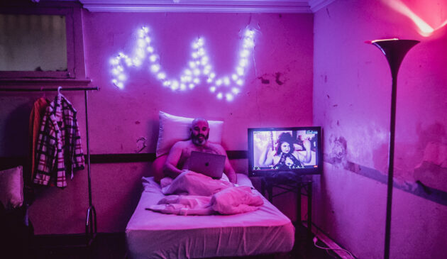 In a dark bedroom lit by pink festoon lights, a woman performs on screen while a shirtless man on a bed watches a laptop