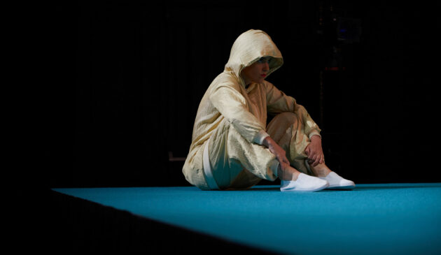 Against a dark background, a person in a pae, loose-fitting hooded tracksuit sits up, knees bent to their chest, on a section of blue floor.
